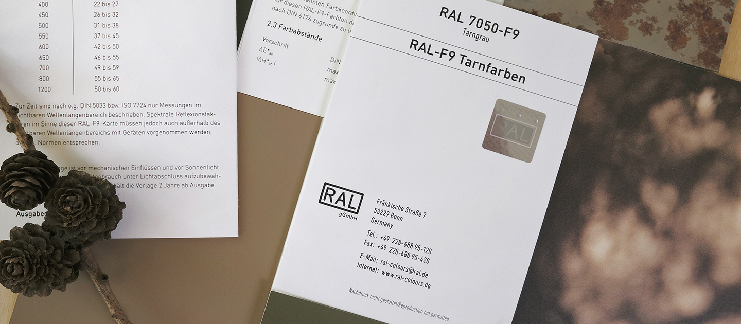 RAL F9 Farbregisterkarte zu den Bundeswehr Tarnfarben. Auf dem Bild liegt mittig, schräg, zugefaltet die Farbregisterkarte zur Farbe "Tarngrau". Im Hintergrund sind weitere Tarnfarben und Tarnmuster zu sehen. In der unteren linken Ecke liegt ein Ast mit Tannenzapfen. | RAL FARBEN

RAL F9 Colour primary standard card for the Bundeswehr camouflage colours. In the picture, the colour sample for the colour "camouflage grey" is folded in the middle, diagonally. More camouflage colours and patterns can be seen in the background. In the lower left corner there is a branch with pine cones. | RAL COLOURS