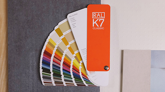 Der RAL K7 Farbfächer liegt aufgefächert auf einem dunkelgrauen Untergrund. Rechts daneben sind verschiedene helle Muster zu sehen. | RAL FARBEN

The RAL K7 colour fan is fanned out on a dark gray background. Various bright patterns can be seen to the right. | RAL COLOURS