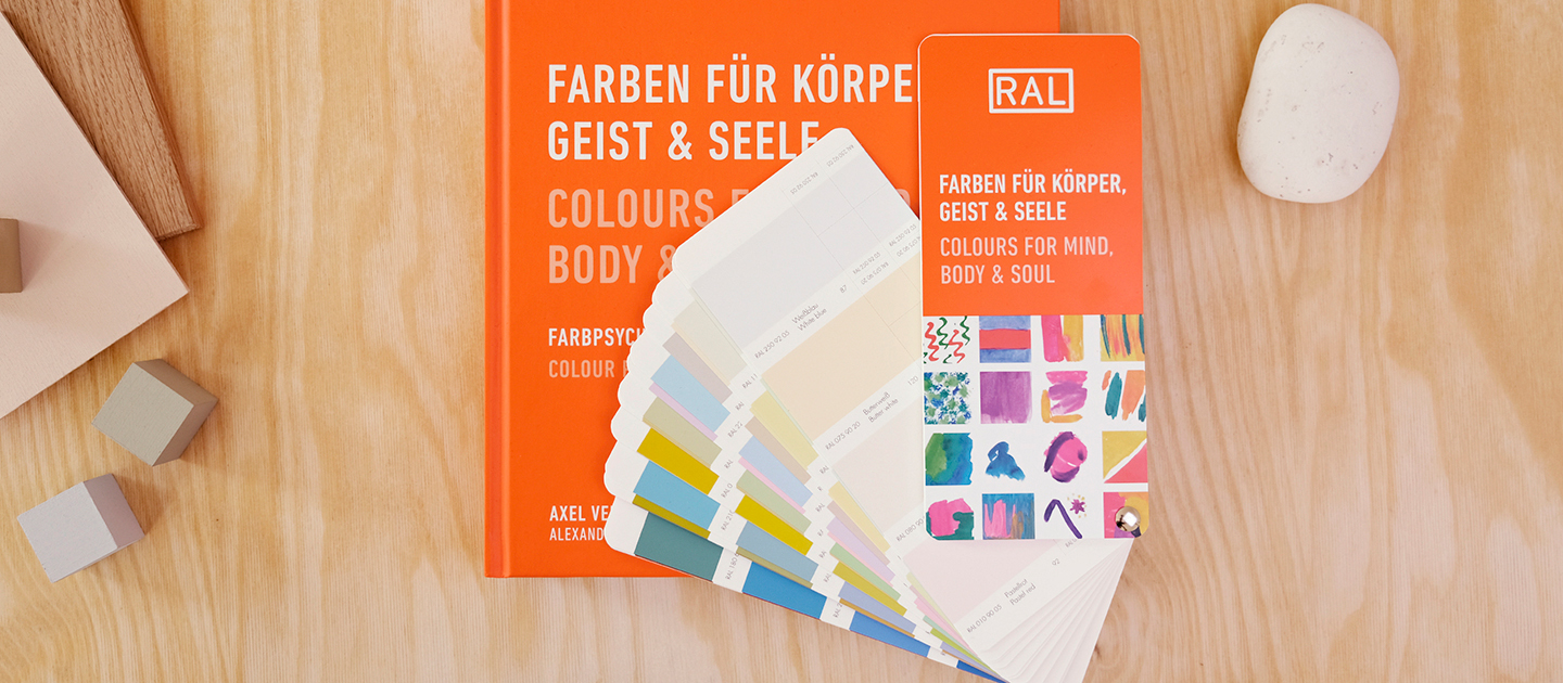 RAL Buch und Farbfächer Bundle: Farben für Körper, Geist & Seele. Das orangene Buch liegt geschlossen auf einem hellen Holzuntergrund, sodass die Aufschrift des Buchtitels zu lesen ist. Auf dem Buch liegt geöffnet der zugehörige Farbfächer mit heraustrennbaren Farbfeldern. Neben dem Bundle liegt rechts ein Stein und links ein paar Holzwürfel. | RAL FARBEN

RAL book and colour fan bundle: Colours for body, mind & soul. The orange book lies closed on a light wooden background so that the title of the book can be read. The corresponding colour fan with removable colour fields lies open on the book. Next to the bundle there is a stone on the right and a few wooden cubes on the left. | RAL COLOURS