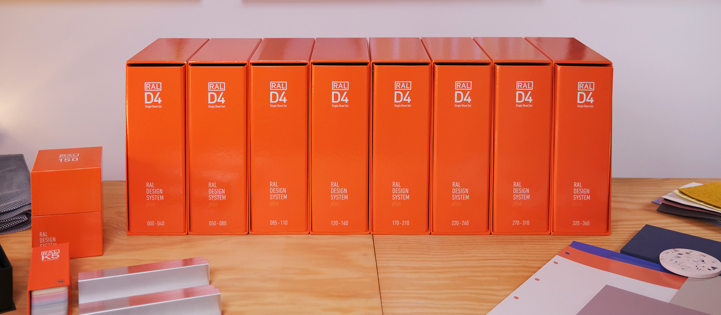 Das RAL D4 Einzelbogen Set A4 mit 8 Ringordnern steht in einem Büro. Daneben liegt links  ein RAL K5 Farbfächer und ein RAL Starter Kit. rechts sind einige RAL D4 Einzelbogen mit weiteren Materialien zu sehen. | RAL FARBEN

The RAL D4 Single sheet set A4 with 8 ring binders is in an office. Next to it on the left is a RAL K5 colour fan and a RAL starter kit. On the right, you can see some RAL D4 Single sheets with other materials. | RAL COLOURS